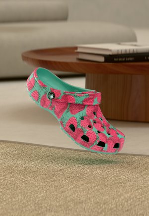 Τυρκουάζ ξύλινο παπούτσι με φωτεινό ροζ σχέδιο φράουλας αιωρούμενο πάνω από μια μπεζ μοκέτα, με ένα ξύλινο τραπέζι και βιβλία στο φόντο.