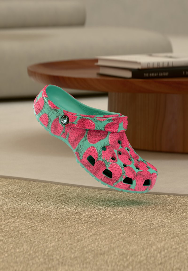 Sabot turquoise avec un motif de fraises rose vif flottant au-dessus d'un tapis beige, avec une table en bois et des livres en arrière-plan.