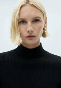 Maglione nero a coste con collo alto, indossato da una modella con capelli corti e biondo chiaro e accessori minimi.