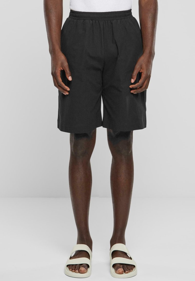 Homme portant un short noir jusqu'aux genoux et des sandales blanches, debout devant un fond clair uni.