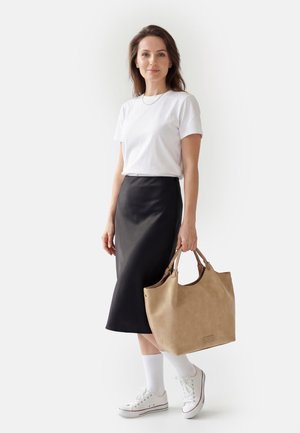 Woman wearing white t-shirt, black midi skirt, white sneakers, and socks, holding a large beige handbag against a plain light background.