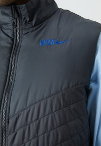 Ciemnoszary, pikowany kamizelka z wysokim kołnierzem, z niebieskimi logo "INTER" i Nike. Teksturowany materiał z poziomymi detalami szycia.