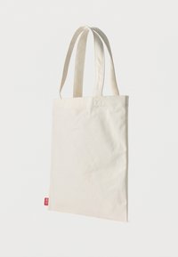 Βαμβακερή τσάντα ώμου σε εκρού χρώμα με δύο μακριές λαβές και κόκκινη ετικέτα μάρκας. Λεία υφή, ορθογώνιο σχήμα και άνετη, ακατάστατη σχεδίαση.