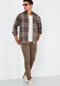 Bruine flanellen overhemd met een een geruit patroon, gecombineerd met beige skinny broek en witte sneakers. Inclusief zonnebril en een gelaagde look.