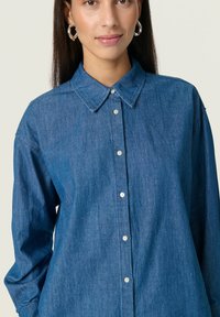 Cămașă din denim cu guler clasic, închidere cu nasturi, mâneci lungi și croi lejer, într-o nuanță de albastru mediu cu o textură moale.