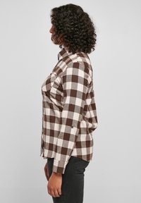 Femme aux cheveux bouclés portant une chemise à manches longues à carreaux marron et blanc et un pantalon foncé, debout de profil sur un fond uni.