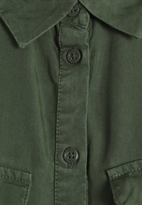 Olivengrøn skjorte med krave, knaplukning og brystlommer. Stoffet har en glat tekstur og en holdbar finish.