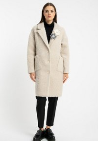 Beżowy płaszcz z wełny z dodatkiem miękkiego włókna o teksturowanym wykończeniu, z dwiema przednimi kieszeniami i trzema guzikami. Kwiatowa broszka jako akcent dodaje stylu. Zestawiony z czarnymi spodniami.
