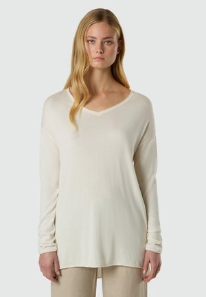 Giovane donna con lunghi capelli biondi che indossa una maglia larga a maniche lunghe color panna con scollo a V e pantaloni beige su uno sfondo semplice.