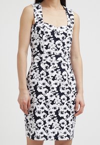 Ärmlös svart och vit blommig klänning med fyrkantig halsringning, figursydd midja och knälång fåll, som bärs av en stående person.