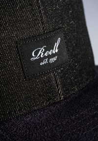 Gorra negra con tejido texturizado, que presenta una etiqueta rectangular prominente con bordado blanco que dice "Reell est. 1997." Parte inferior azul oscura.
