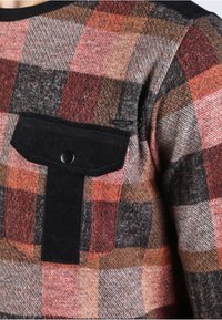 Karierte Hemd mit einer Mischung aus Rot, Orange, Schwarz und Grau. Enthält eine schwarze Cord-Tasche auf der Brust mit einem Druckknopf.