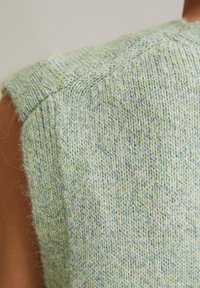 Gilet tricoté vert clair avec une surface texturée, présentant des éclats bleus et un détail de couture à l'épaule. Tissu en mélange de laine douce.