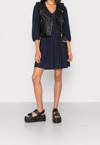 Un gilet de moto en cuir noir porté sur une robe bleu foncé avec un motif texturé, associé à des sandales noires épaisses avec une semelle épaisse.