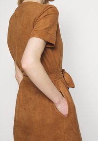 Robe en daim marron à manches courtes, taille ajustée et poches latérales. Comprend un détail de ceinture nouée à la taille pour plus de définition.
