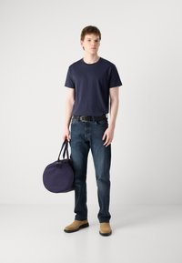 T-shirt en coton marine, jeans bleu foncé, chaussures beiges, tenant un sac rond marine avec deux poignées. Design simple, apparence décontractée, arrière-plan neutre.