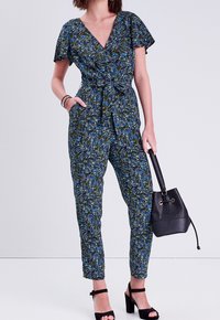 Jumpsuit à motif floral vert, bleu et noir ; décolleté en V, manches courtes, taille cintrée, jambes fuselées et poches. Sac noir et talons.