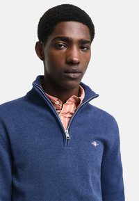 Maglione zip-up blu navy realizzato in tessuto a maglia, con colletto alto e un piccolo logo. Indossato sopra una camicia a righe rosa chiaro con colletto.