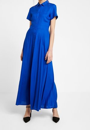 Vrouw draagt een felblauwe, korte mouwen, knopsluitende maxi-jurk gecombineerd met zwarte pumps met spitse neus, staand tegen een witte achtergrond.