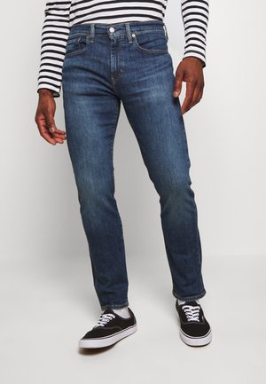Jean en denim bleu foncé avec une coupe slim, cinq poches et des coutures contrastées, associés à des baskets noires et une chemise à rayures à manches longues.