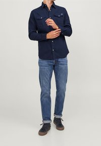 Camicia navy con bottoni, due tasche frontali, maniche lunghe e bottoni metallici a contrasto. Indossata con jeans azzurri arrotolati e sneakers scure.