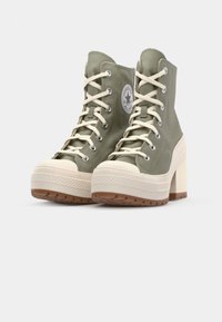 Zapatillas altas de lona verdes con cordones blancos, puntera de goma y tacón grueso color crema; incluye un parche redondo con el logo en el lateral.