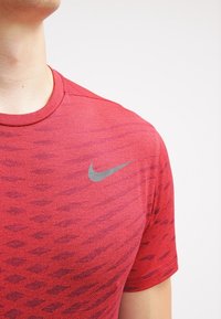 Camiseta deportiva roja con un patrón texturizado y mangas cortas. Presenta un logo de Nike en gris en la parte superior izquierda.