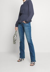 Mörkblå peplumblus med långa ärmar, kombinerad med blå utsvängda jeans. En svart-och-vitt zebramönstrad väska i handen; öppetåiga klacksandaler.