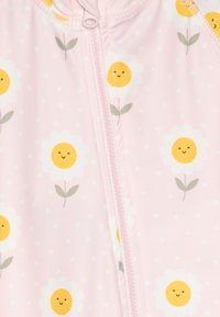 Lyserød zip-up heldragt med hvide blomster med gule midterpunkter og grønne blade, prikket mønster på et blødt stof. Lynlåslukning foran.