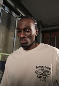Camiseta oversized beige con un gráfico negro de un tanque y el texto "INFAMOUS GRMY". Material de algodón con una textura suave.