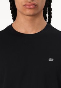 T-shirt en coton noir avec un col rond et des manches courtes. Présente un petit logo "GAP" blanc sur la zone gauche de la poitrine.
