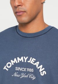 Mėlynas medvilninis sportinis džemperis su apvalia iškirpte, iš priekinės pusės užsiūta balta emblema "TOMMY JEANS" ir "SINCE 1985".