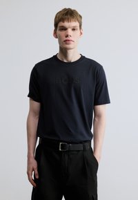 Tengerészkék pamut rövid ujjú póló tónusos "BOSS" logóval. Fekete nadrággal és hozzáillő övvel viselve, minimalista dizájn.