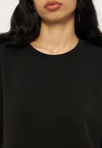 Femme aux longs cheveux bruns porte un t-shirt noir uni à col rond et des boucles d'oreilles fines en chaîne, lèvres fermées et expression neutre.