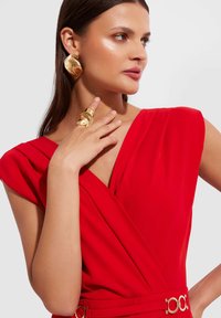 Czerwona sukienka w stylu owijanym z krótkimi, luźnymi rękawami. Posiada złoty pasek akcentowy oraz dużą biżuterię w złotym kolorze, podkreślającą gładką fakturę materiału.