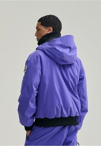 Lila puffertöltött dzseki kapucnival, bordás fekete szegéllyel és oldalsó zseb részletekkel. Simas texture és laza fazon.