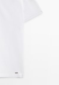 T-shirt blanc à manches courtes en coton avec une texture lisse, présentant un design minimaliste et un petit logo près de l'ourlet.