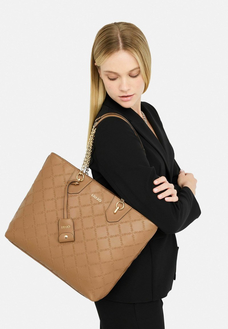 Torebka tote w kolorze tan, strukturalna, z wytłaczanym wzorem, łańcuchem i skórzanymi ramiączkami, z kluczowym akcentem. Zaprezentowana na modelce w czarnym żakiecie.
