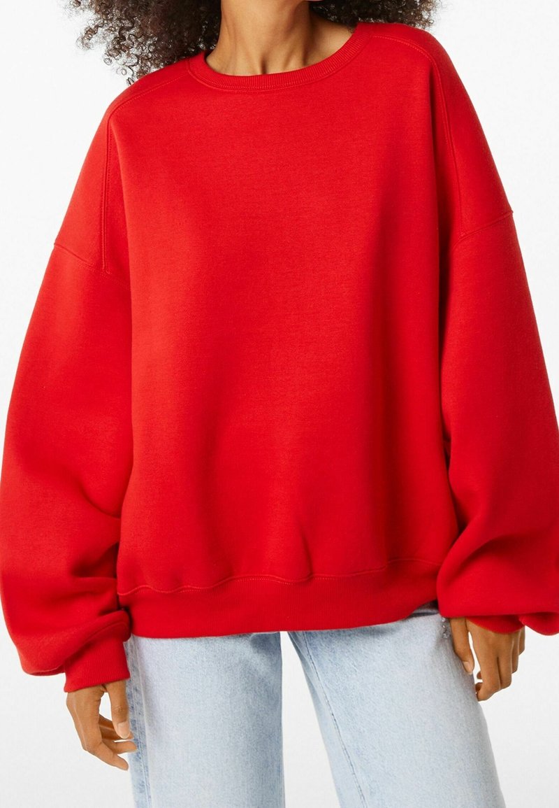 Sweatshirt rouge surdimensionné avec épaules tombantes, encolure ronde côtelée et manches resserrées aux poignets. Fabriqué en tissu doux et texturé. Associé à un jean bleu clair.