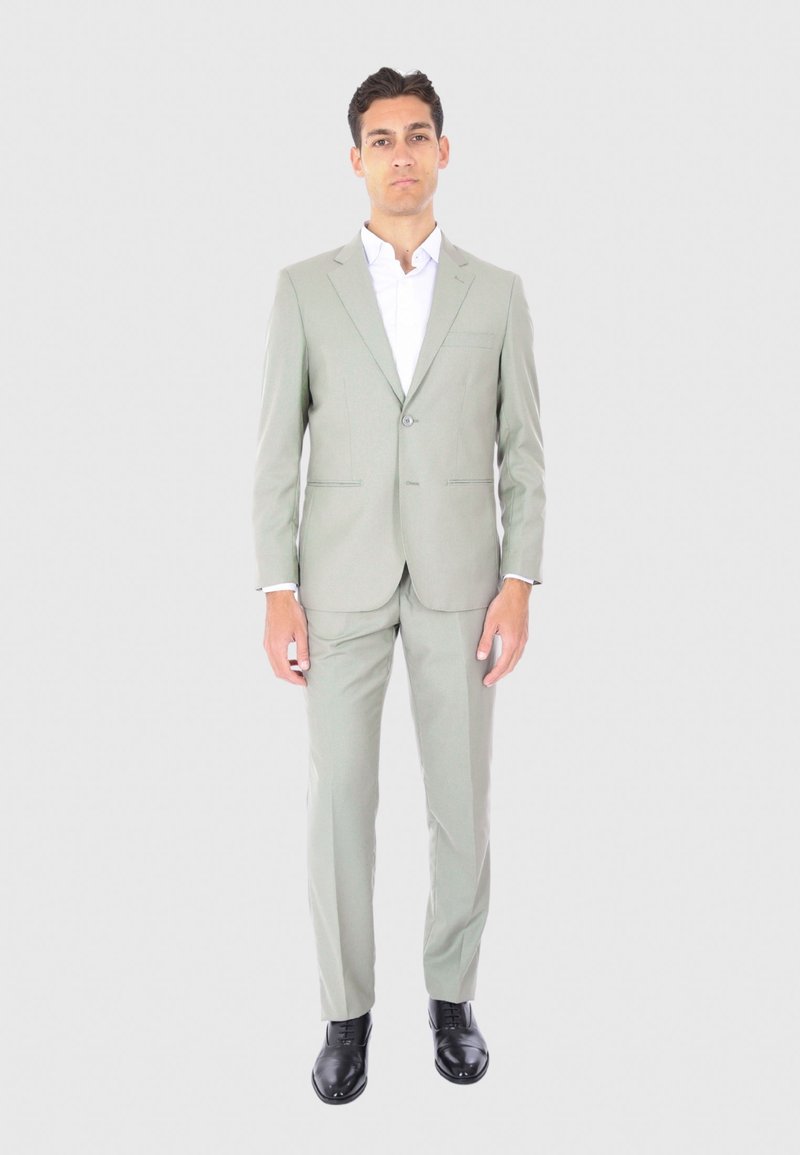 Abito verde chiaro con chiusura a un bottone, rever a punta e pantaloni dritti. Abbinato a una camicia bianca e scarpe nere lucide.