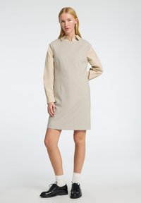 Beige šaty typu shift s krátkými rukávy, s límečkovým výstřihem a hladkým povrchem. V kombinaci s černými botami a bílými ponožkami.