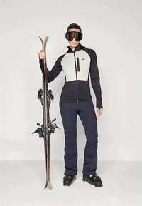 Ski outfit featuring a black and light gray zip-up jacket, dark ski pants, and black ski boots, holding skis and wearing large goggles.