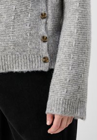 Cardigan lavorato a maglia grigio chiaro con un motivo a trama, dotato di quattro bottoni marroni, polsini a costine e un orlo leggermente sfilacciato.