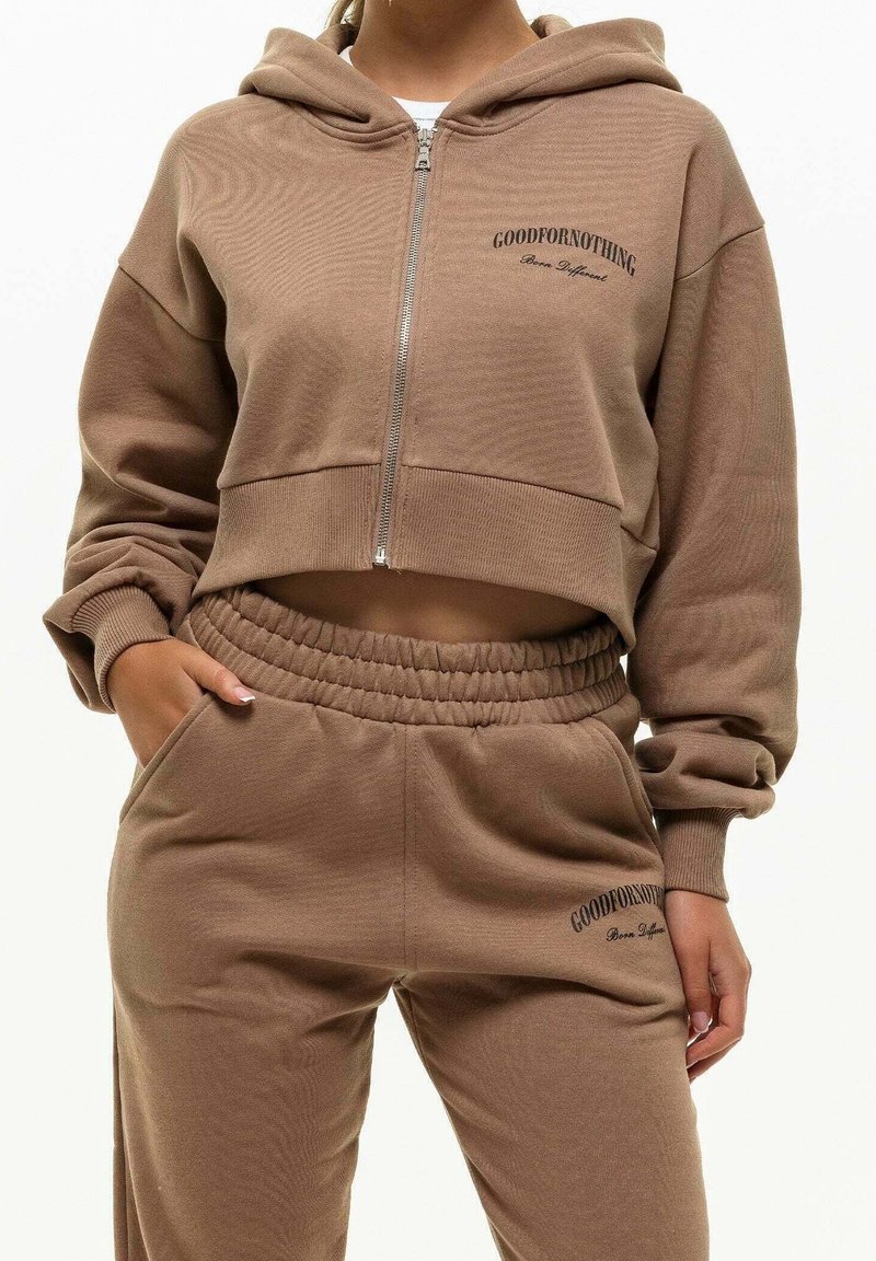 Oříznutá hnědá mikina s kapucí na zip a vyšitým logem, doplněná o ladící zúžené tepláky s elastickým pasem a kapsami.
