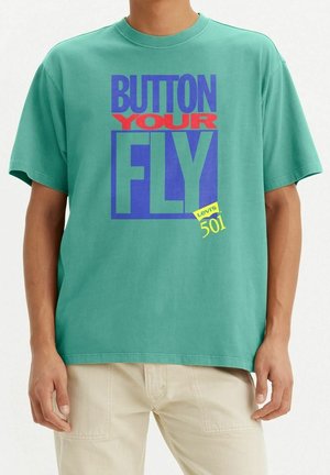 T-shirt en coton vert arborant un texte graphique audacieux en rouge, bleu et jaune. Coupe décontractée avec manches courtes et encolure ronde.