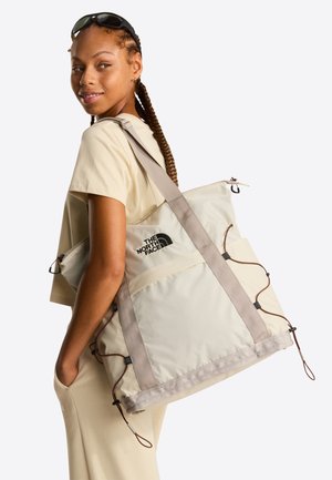 Sac fourre-tout beige avec des bretelles grises, arborant un logo, des cordons de serrage pour la fermeture et un tissu texturé.