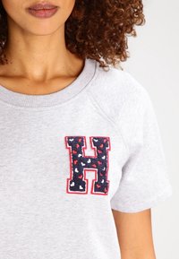 Grå kortärmad sweatshirt med en broderad "H" i marinblått och rött som har ett hjärtmönster. Rund hals och mjuk textur.