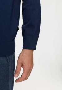 Pulover tricotat de culoare albastru marin, cu manșete ribbate, având o etichetă mică neagră pe partea stângă. Prim-plan al unei mâini odihnindu-se pe lateral.