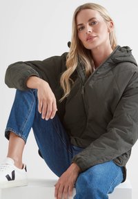Blonde woman sitting with one knee up, wearing a dark green quilted jacket, blue jeans, and white sneakers with black accents.
