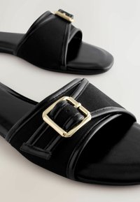 Sandales noires à enfiler avec bouts carrés et larges brides ornées de boucles rectangulaires dorées.